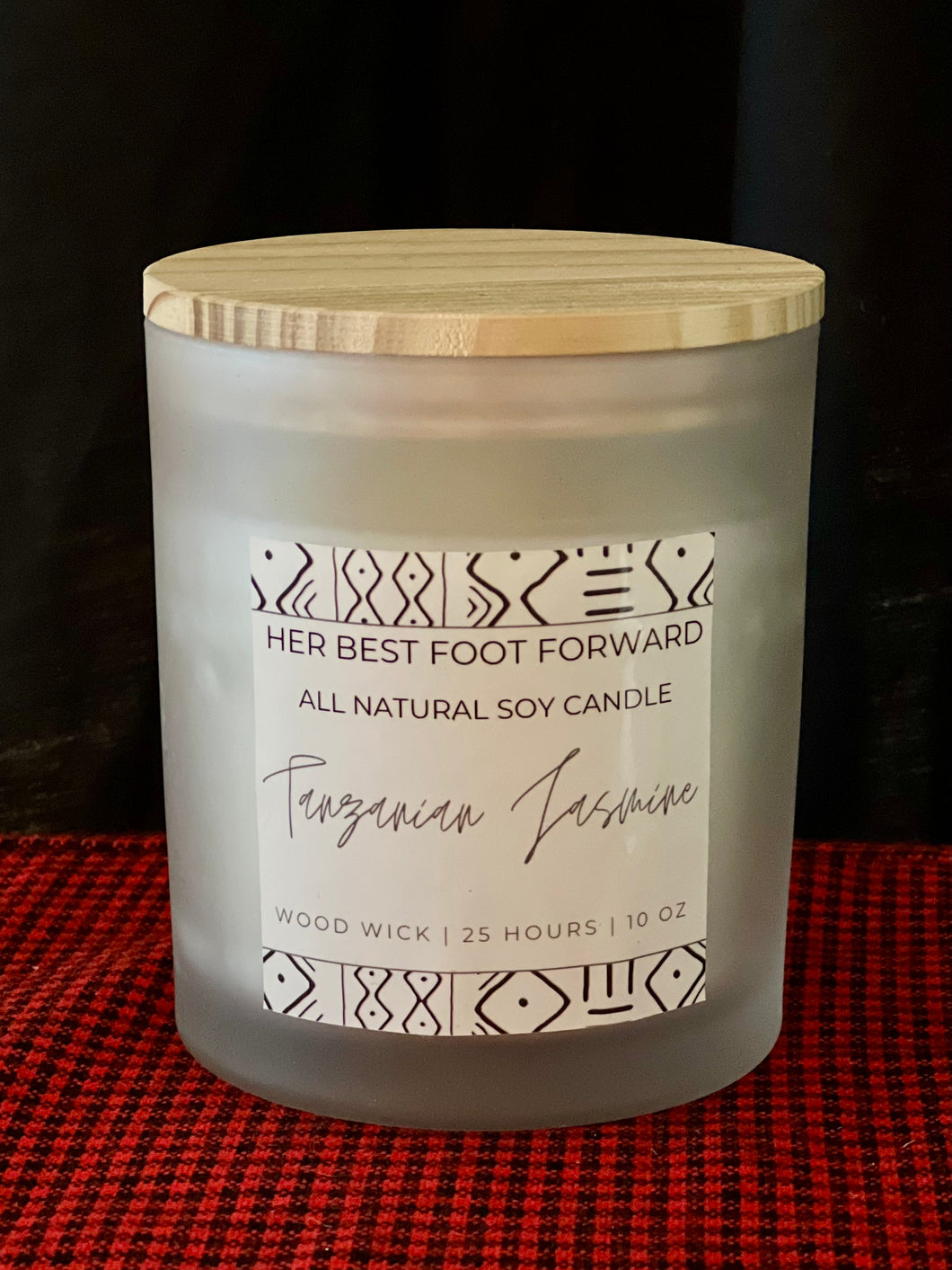 A frosted glass soy wax candle with a wooden lid sits on red and black fabric. The label reads Her Best Foot Forward You GLOW Girl Soy Wax Candle, Natural Wood Wick, African-inspired scents, 10 oz, 25 hours burn time.