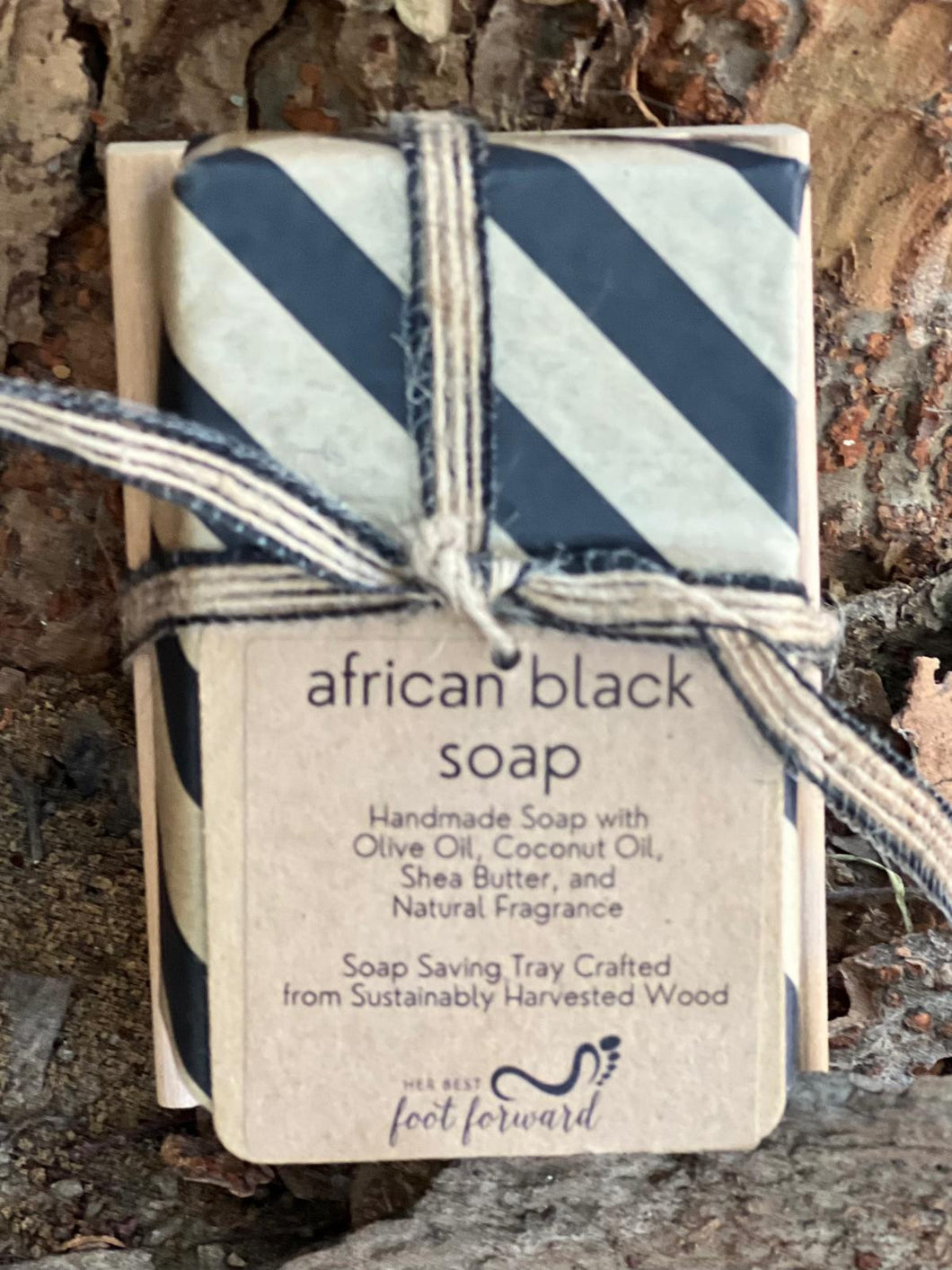 A Handcrafted Soap Set with Wooden Tray by Her Best Foot Forward, featuring natural African-inspired scents, rests on a tree bark background. Wrapped in striped paper and twine for an artisanal touch.